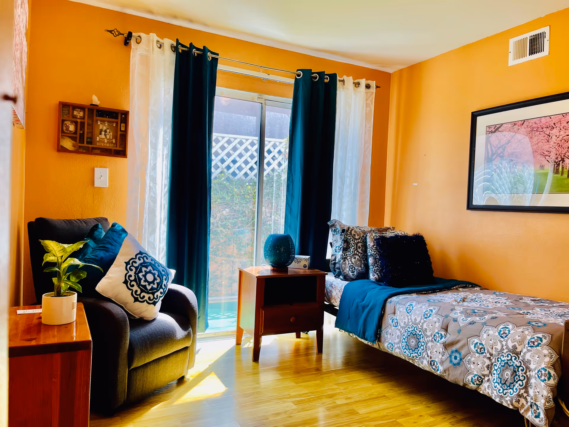 A cozy bedroom with orange walls featuring a single bed with patterned bedding and multiple pillows. Next to the bed is a wooden nightstand with a decorative vase and a box of tissues. A comfortable armchair with decorative pillows and a small potted plant on a wooden side table are positioned near a sliding glass door with dark teal and white curtains. A framed picture of pink cherry blossoms hangs on the wall.