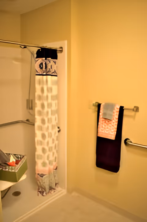 A bathroom with a shower area featuring a curtain with a patterned design. There is a towel rack on the wall holding three towels in different colors, and a grab bar is installed nearby. A small shelf with a green box is visible near the shower.