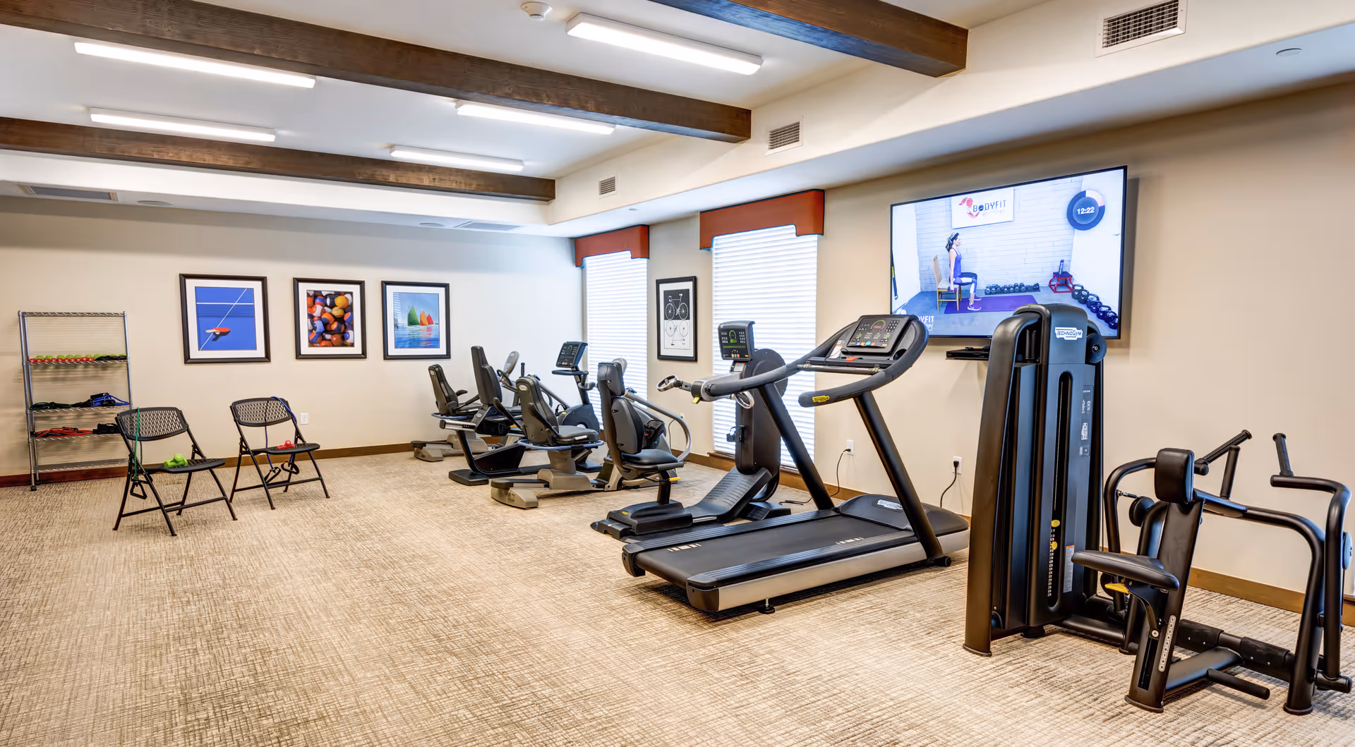 A senior living facility fitness room with exercise equipment including treadmills, stationary bikes, and strength machines. The room has beige carpet, white walls with framed artwork, two windows with blinds, and a large TV mounted on the wall displaying an exercise video. Two folding chairs and a rack with small weights are also visible.