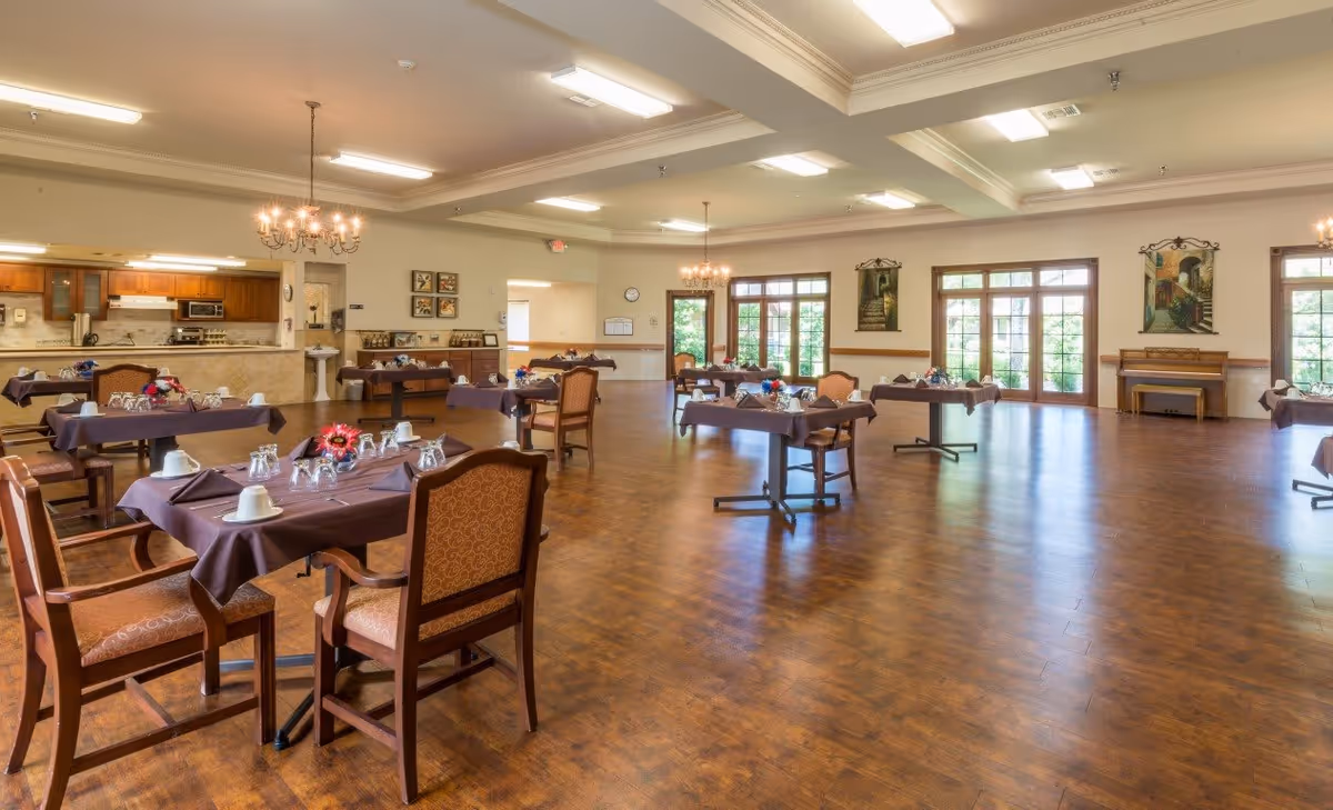 Spacious dining room with multiple tables set with place settings, chandeliers, and large windows overlooking greenery.