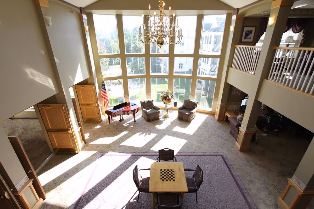Bright and spacious living area with large floor-to-ceiling windows letting in natural light. The room features a chandelier hanging from the ceiling, a small table with a chessboard and four chairs on a rug, two armchairs near a floral arrangement, and a grand piano with an American flag beside it. The space has a high ceiling and an upper balcony with white railings overlooking the area.
