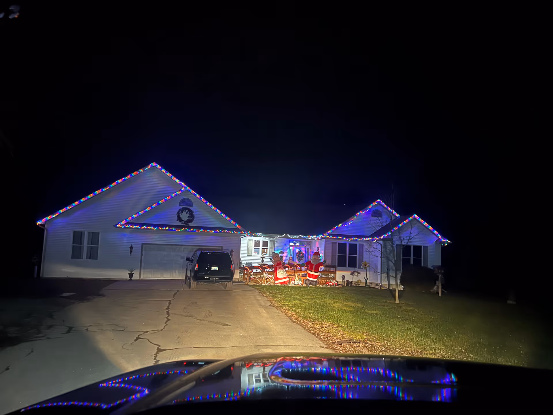 Night view of a single-story house decorated with colorful Christmas lights along the roof edges. The driveway leads to a garage with a black vehicle parked in front. The front yard has Christmas decorations including inflatable Santa Claus figures and a wreath on the garage wall.