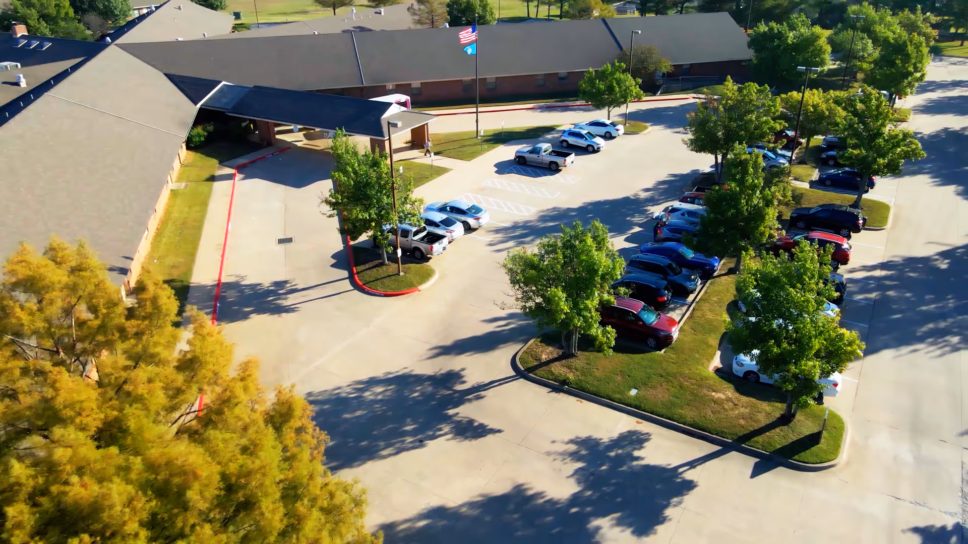 Aerial view of the parking lot and entrance of a senior living facility with several parked cars, green trees, and a building with a covered drop-off area. Two flags are visible near the entrance.