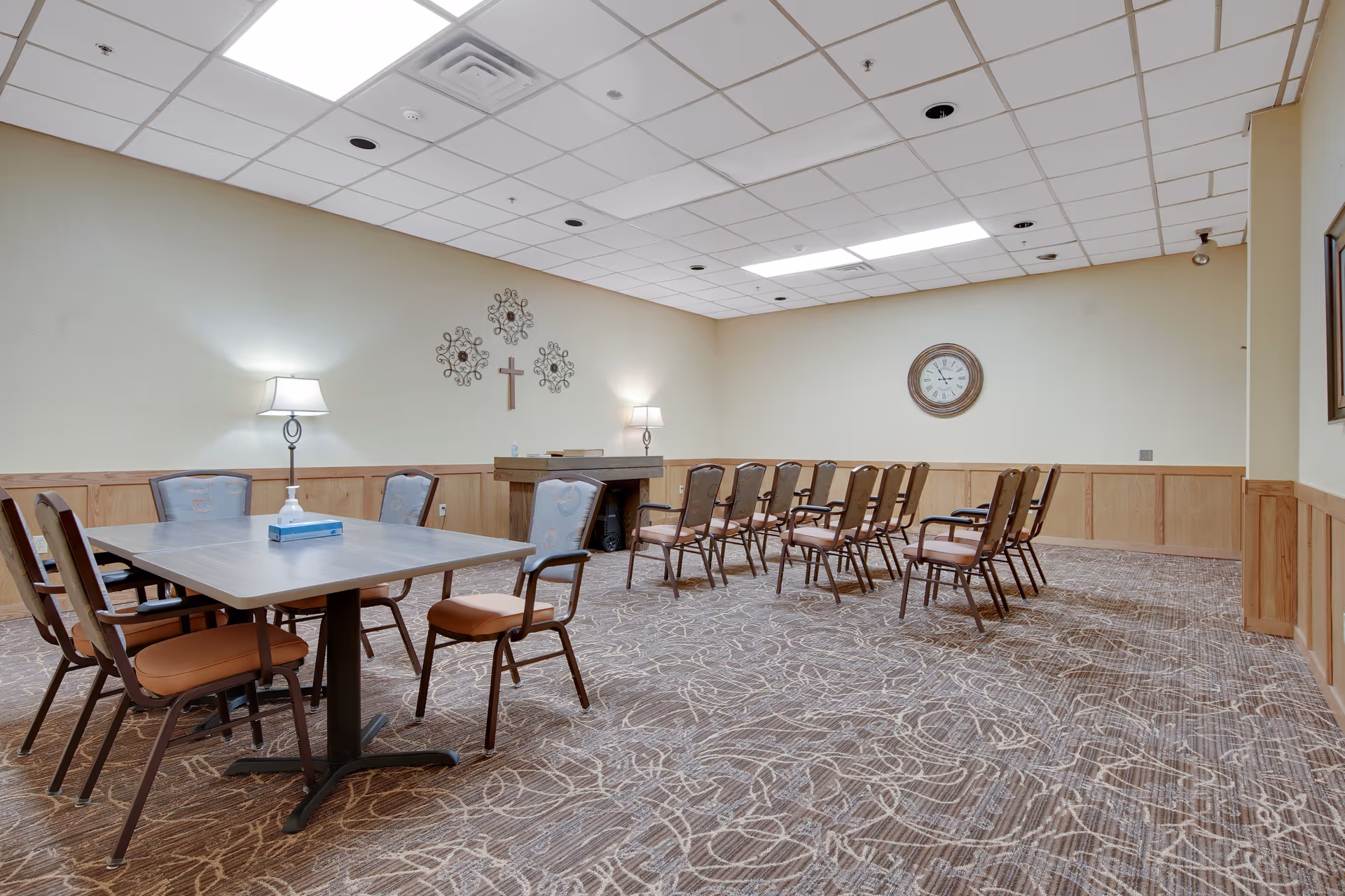 A meeting or activity room with a rectangular table surrounded by chairs on the left and several rows of chairs arranged facing a small podium at the front. The room has beige walls with wood paneling, a patterned carpet, two wall lamps, decorative wall hangings including a cross, and a large clock on the far wall.