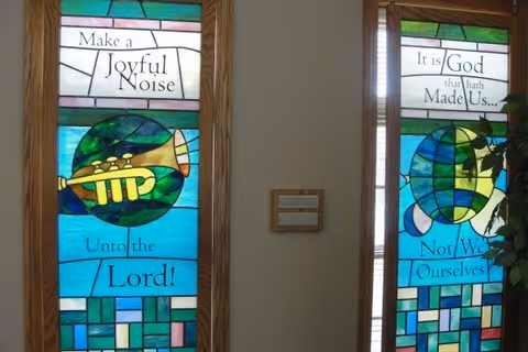 Two vertical stained glass windows with colorful geometric patterns and images of trumpets. The left window reads 'Make a Joyful Noise Unto the Lord!' and the right window reads 'It is God that hath Made Us... Not We Ourselves'. A small plaque is mounted on the wall between the windows.