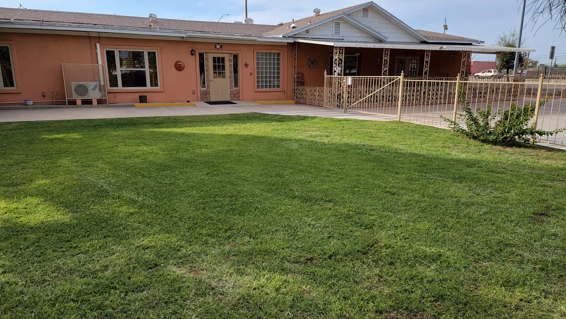 A single-story building with a peach-colored exterior and a white roof, featuring several windows and doors. In front of the building is a well-maintained green lawn and a metal fence enclosing a small porch area. The sky is clear and blue.