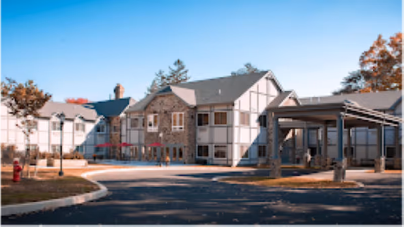 Exterior view of a senior living facility building with Tudor-style architecture, featuring white walls with wooden beams and stone accents. The building has multiple windows and a covered entrance area. There are trees with autumn foliage and a clear blue sky in the background.