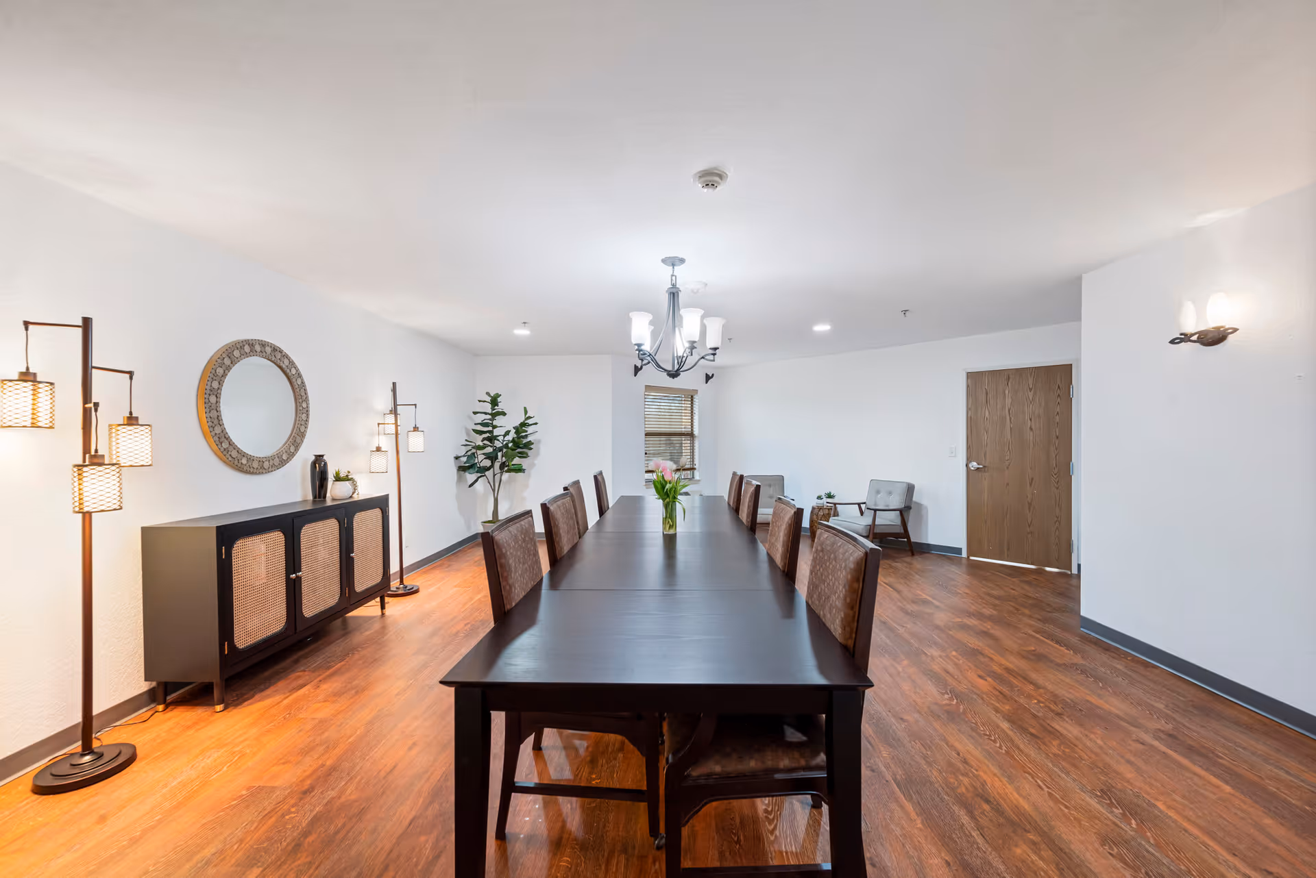 A spacious dining room with a long dark wooden table surrounded by chairs. The room features wooden flooring, a sideboard with decorative items and a round mirror above it, floor lamps, a chandelier, and a small seating area with two chairs near a wooden door.