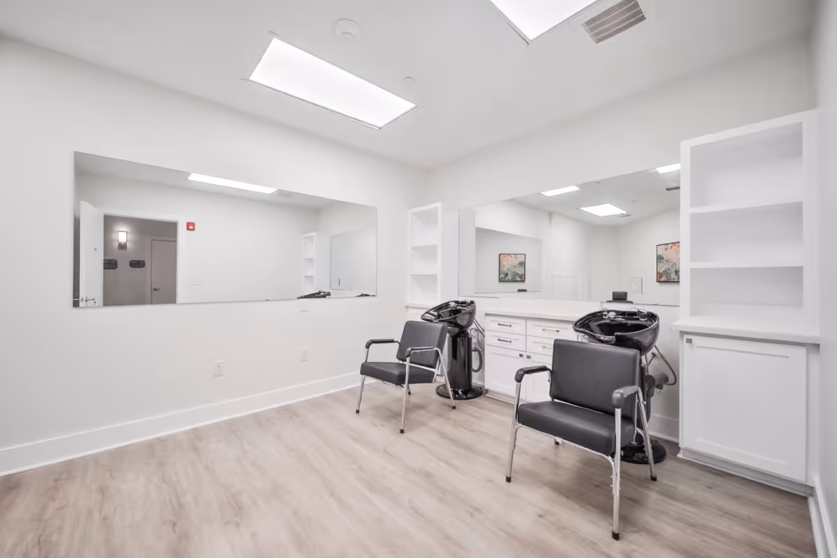 Bright hair salon with two styling chairs, wash basins, large mirrors, and white cabinetry in a senior living facility.
