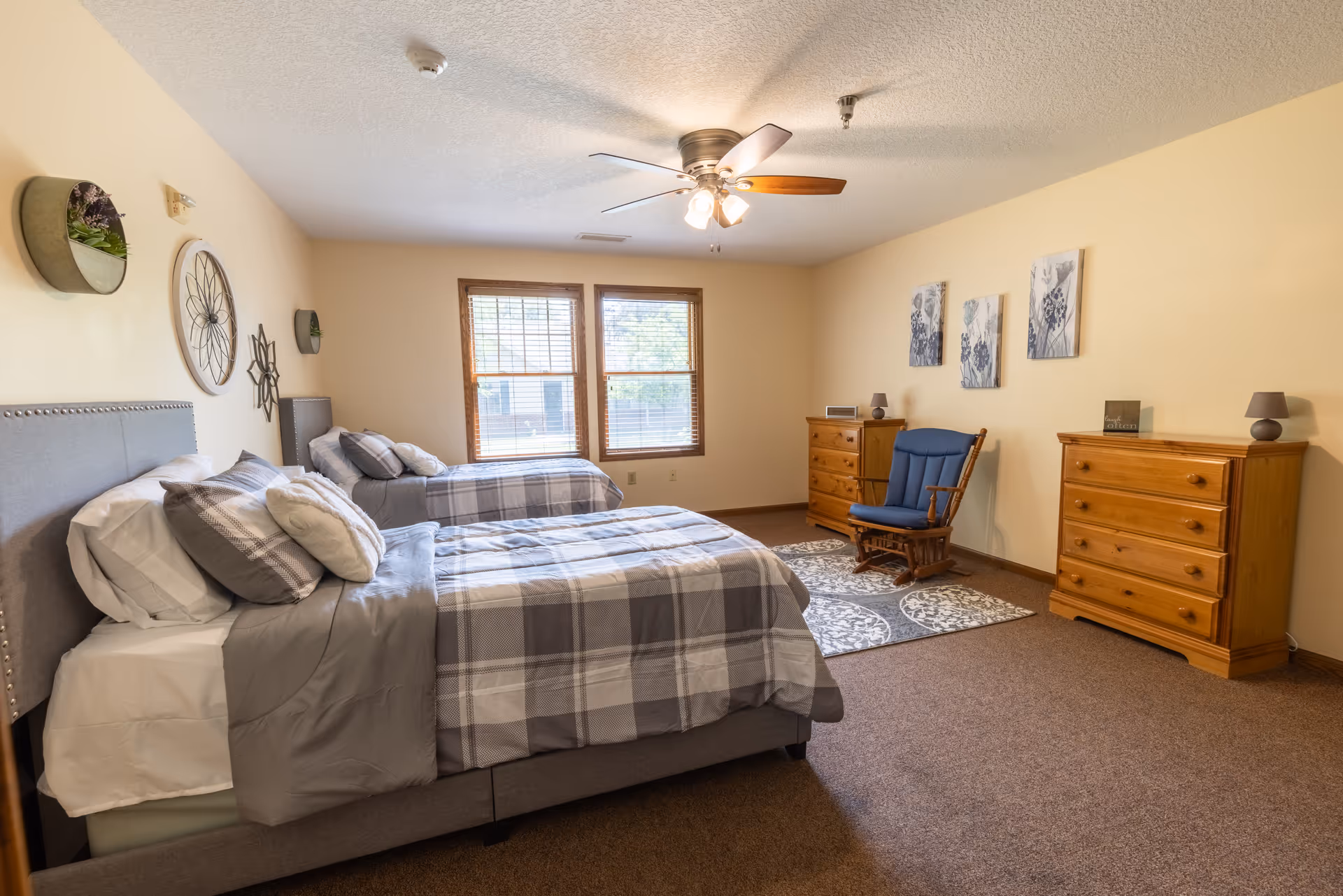 A spacious bedroom with two twin beds dressed in gray and white plaid bedding. The room features beige walls, a ceiling fan with lights, two wooden dressers, a blue cushioned rocking chair on a patterned rug, and three floral wall art pieces above the dressers. Two windows with wooden blinds allow natural light into the room.