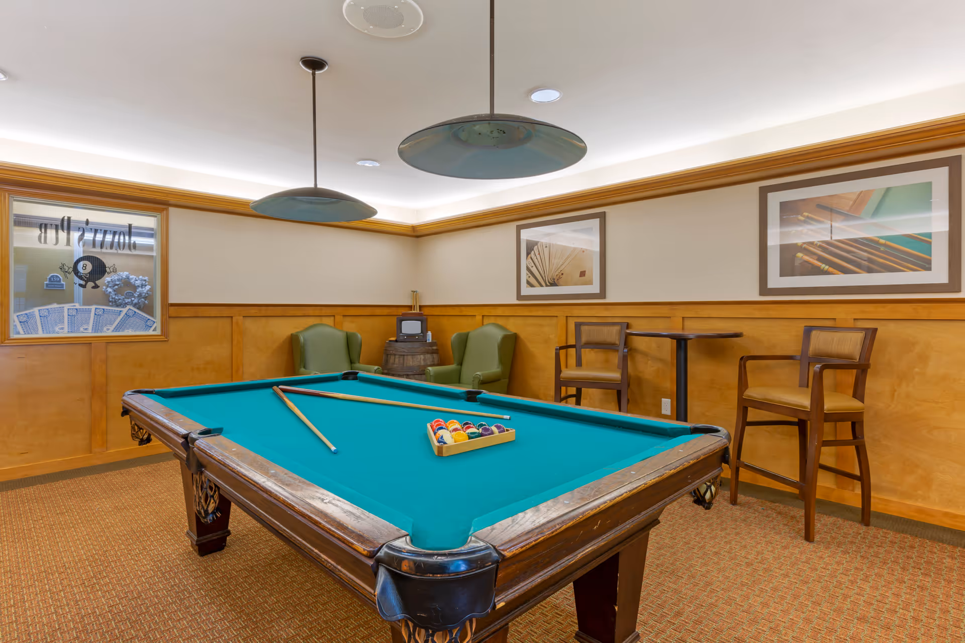 A game room with a pool table in the center, two pool cues resting on the table, and a rack of billiard balls arranged for a game. The room has wood paneling on the walls, two green armchairs, a small round table with two wooden chairs, and framed pictures of playing cards and pool cues on the walls. Two pendant lights hang from the ceiling above the pool table.