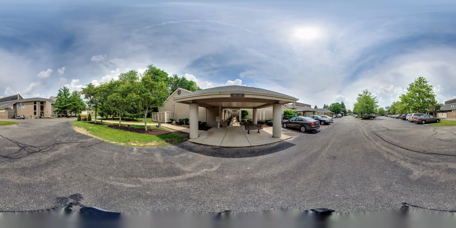 Covered entrance/porte-cochère and surrounding parking lot of a retirement community with cars and trees under a partly cloudy sky.