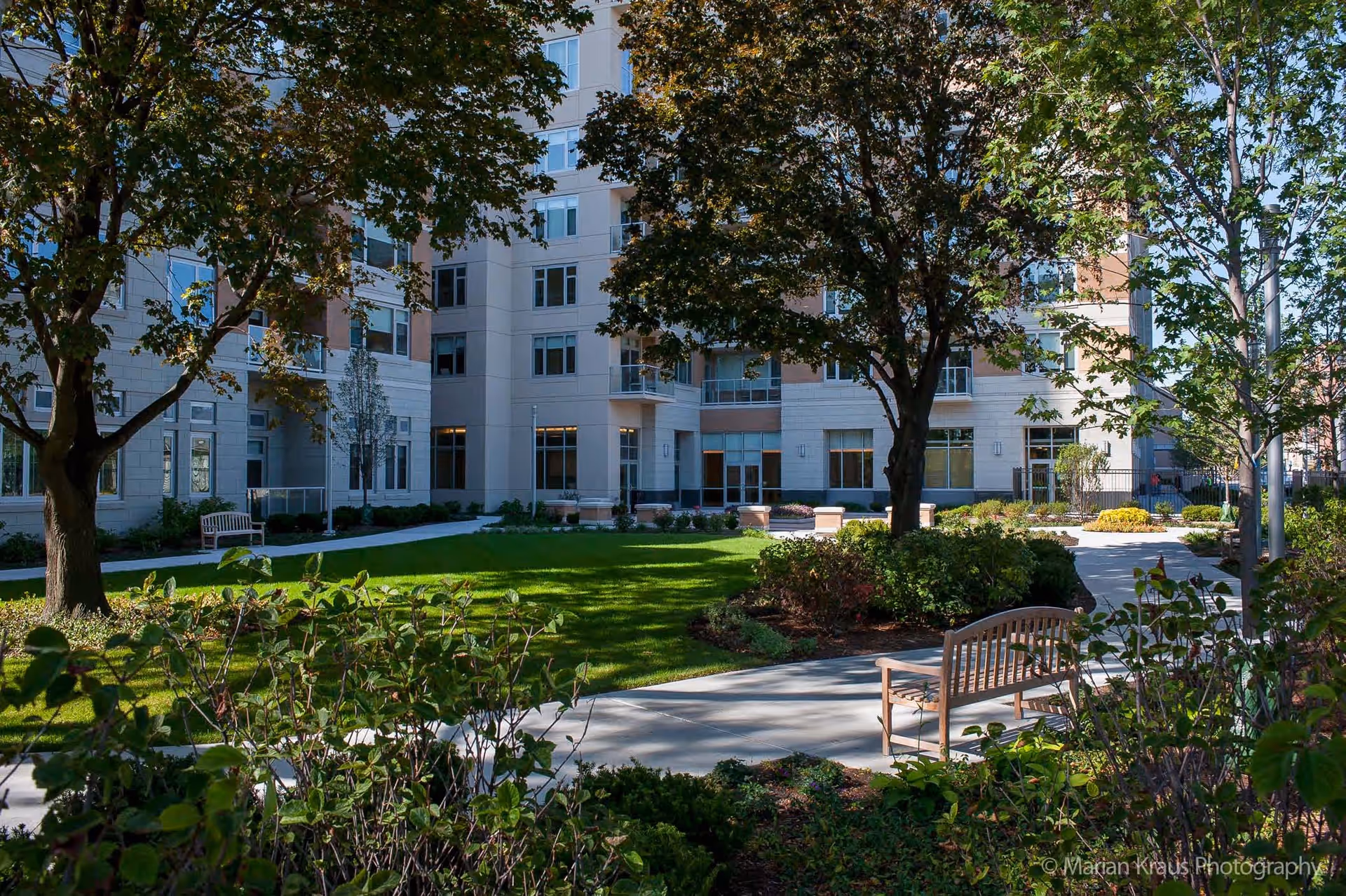 Outdoor garden area with green grass, trees, shrubs, and wooden benches in front of a multi-story residential building under a clear blue sky.