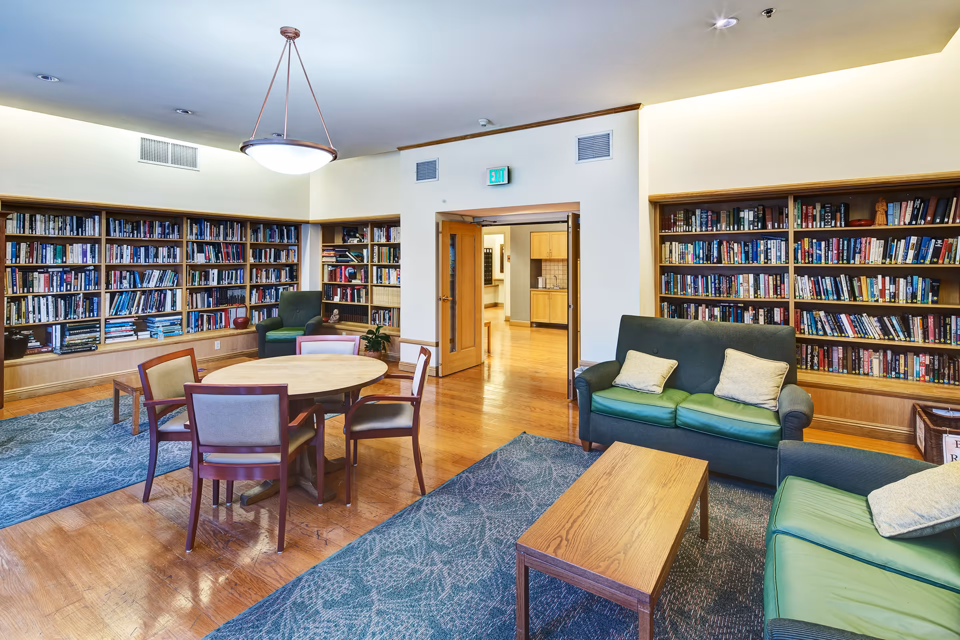 A bright common library/lounge with floor-to-ceiling bookshelves, a round table with chairs, and green sofas around a coffee table.