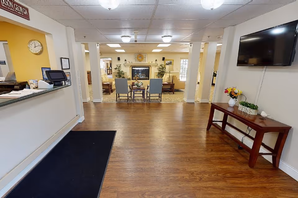 Entrance lobby of a senior living facility with a reception desk to the left, a seating area and fireplace ahead, and a TV on the right.