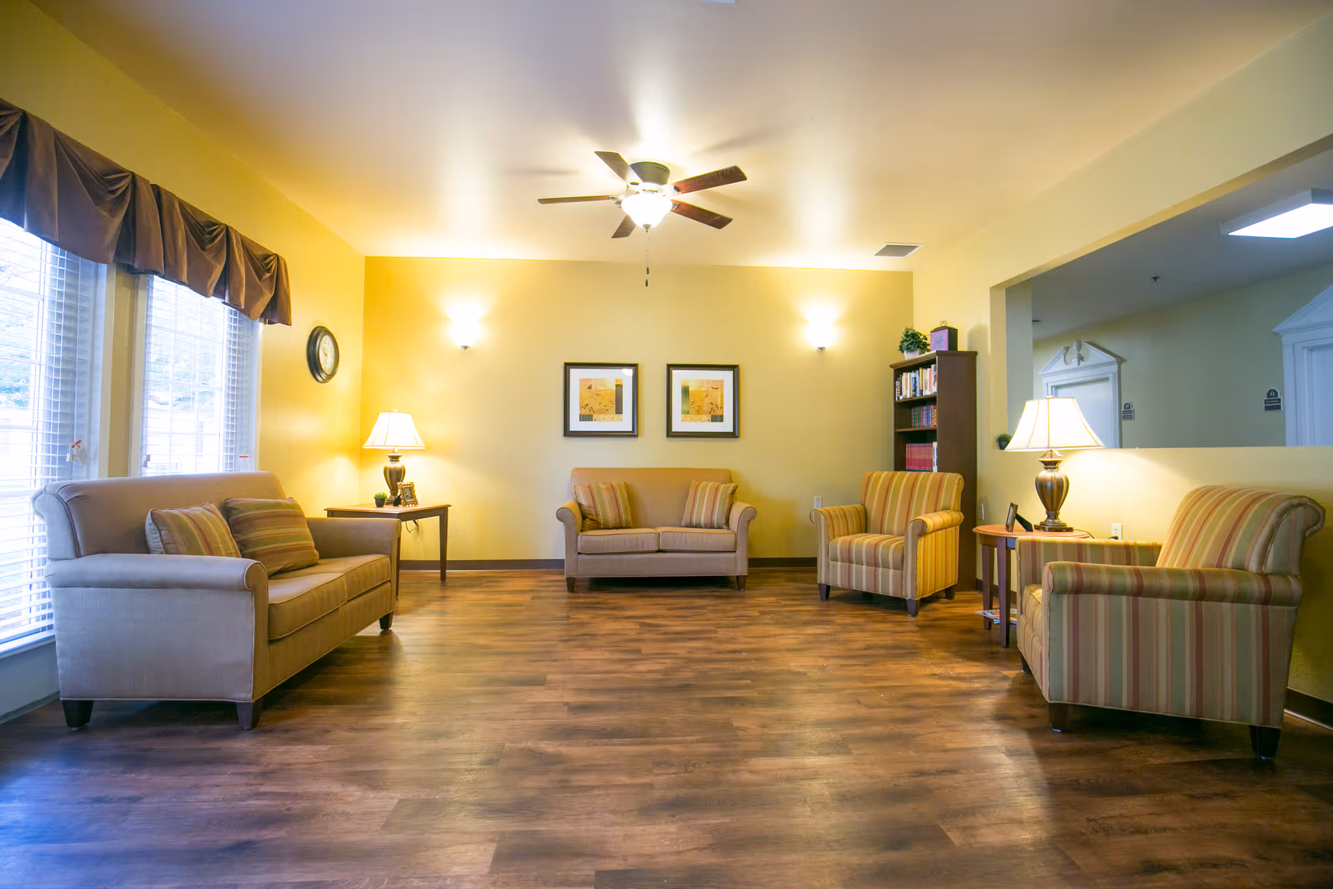 A cozy living room with wooden flooring, yellow walls, and a ceiling fan with light. The room features two beige sofas with striped cushions, two striped armchairs, two side tables with lamps, a bookshelf, two framed pictures on the wall, and large windows with blinds and valances.