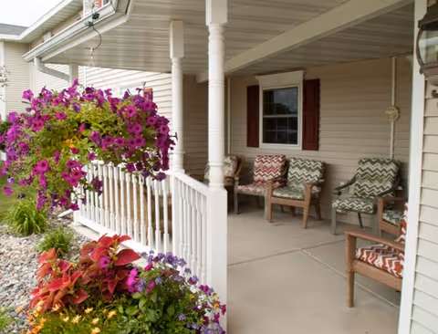 Covered front porch with wicker chairs, hanging baskets and potted flowers beside a railing.