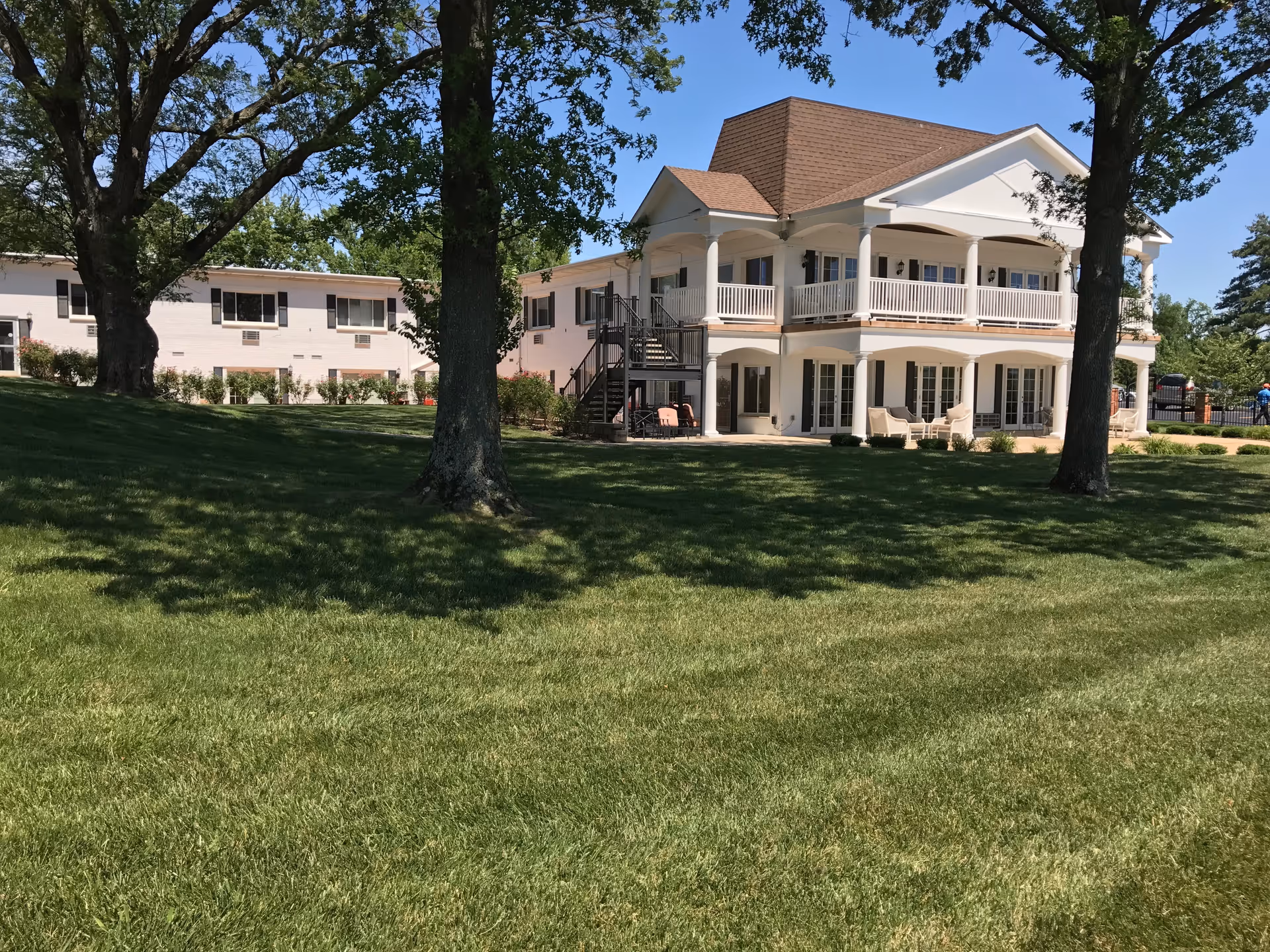 Two-story white nursing home building with wraparound porches, a large grassy lawn, and shade trees.