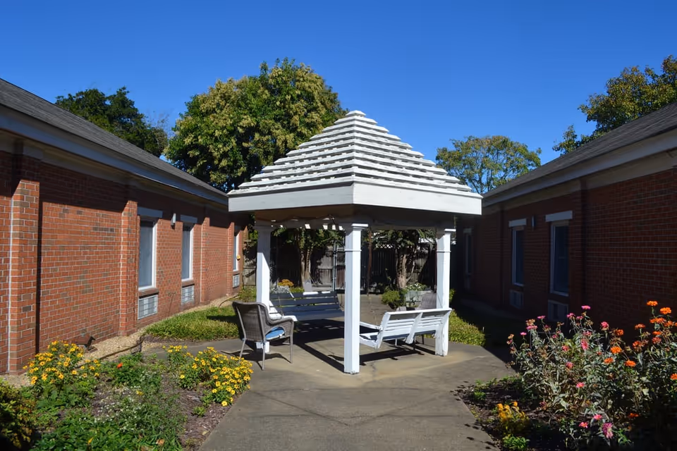 Outdoor courtyard area at Mayfair House Senior Living with a white wooden gazebo featuring a pyramid-shaped roof. The gazebo has benches and chairs underneath. The courtyard is surrounded by red brick buildings and landscaped with colorful flowers and green shrubs under a clear blue sky.