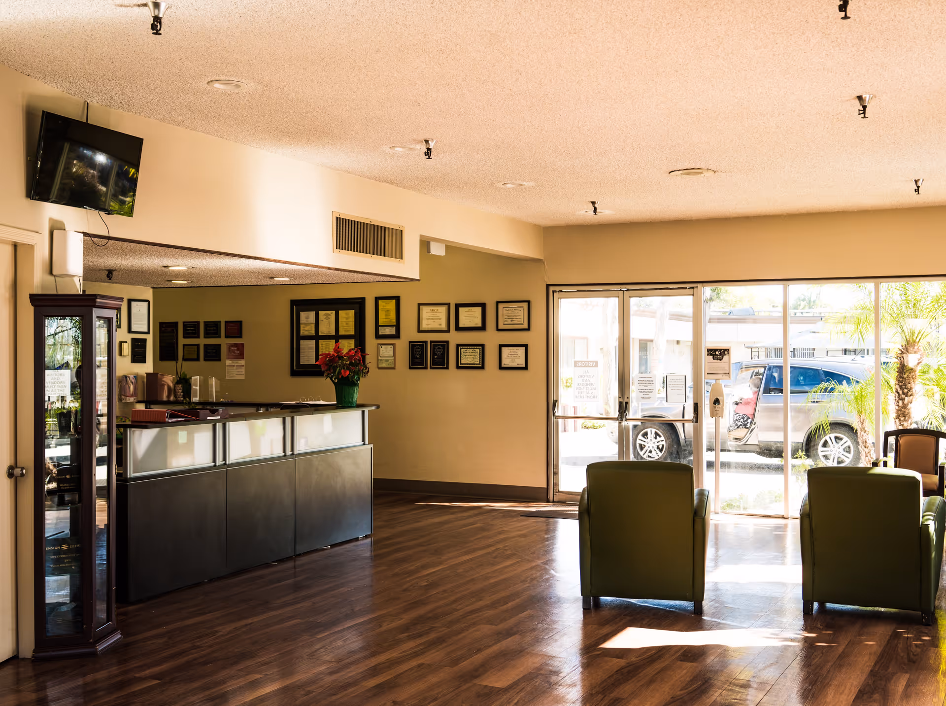 Reception area of Whittier Hills Healthcare Center with a modern front desk, a TV mounted on the wall, framed certificates on the wall behind the desk, and green chairs facing large glass doors that lead outside where a vehicle is parked.