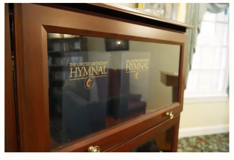 A wooden cabinet with glass doors displaying a book titled 'The United Methodist Hymnal' inside. The room has a window with curtains and a patterned carpet.