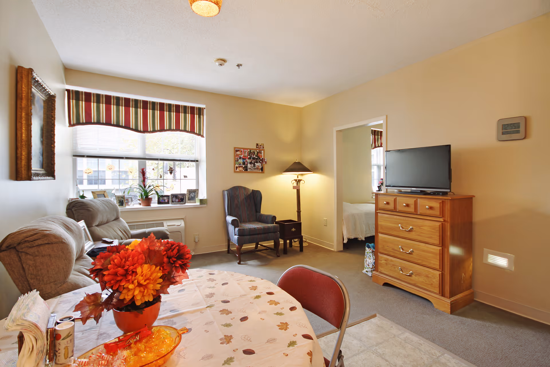 A cozy living room area in a senior living facility with a beige sofa, a striped armchair, a wooden dresser with a TV on top, and a round table covered with a floral tablecloth and a vase of red and orange flowers. A window with striped valance lets in natural light, and a doorway leads to a bedroom with a bed visible.