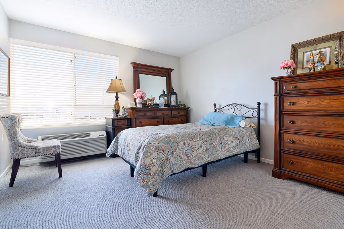 A bright bedroom with a large window covered by white blinds. The room features a bed with a patterned bedspread and blue pillows, a wooden dresser with a mirror, a wooden chest of drawers, a decorative lamp, a floral chair, and various decorative items including flowers and framed pictures.