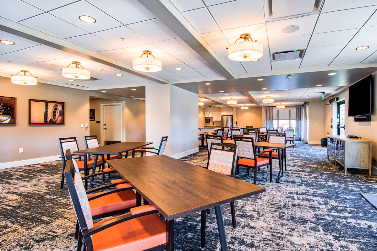 A spacious dining area in Clarendale at Bellevue Place featuring multiple wooden tables with chairs that have orange cushions and patterned backs. The room has a patterned carpet, ceiling lights, framed artwork on the walls, and a large flat-screen TV mounted on the right wall. The back of the room includes a kitchen area with stainless steel appliances and large windows with curtains.