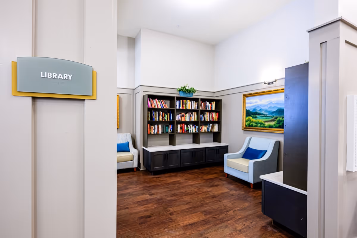 Interior view of a library room in a senior living facility with a bookshelf filled with books, two armchairs with blue cushions, a framed landscape painting on the wall, and a sign on the wall that reads 'LIBRARY'.