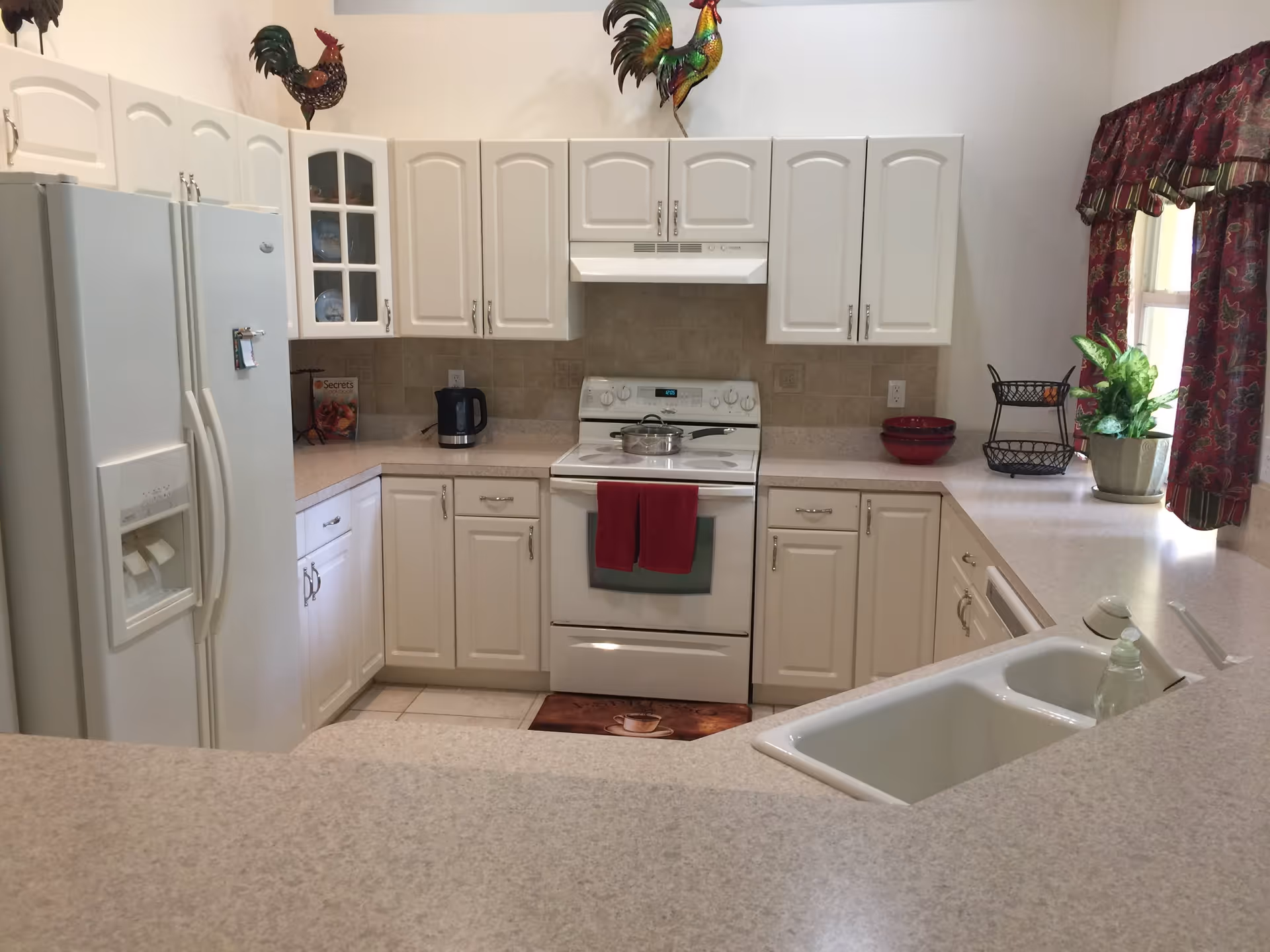 A clean and well-lit kitchen with white cabinets, a white refrigerator, an electric stove with a pot on it, and a double sink. The kitchen has beige countertops and a tiled backsplash. There are decorative rooster figurines on top of the cabinets, a plant near a window with red patterned curtains, and a red towel hanging on the oven door.
