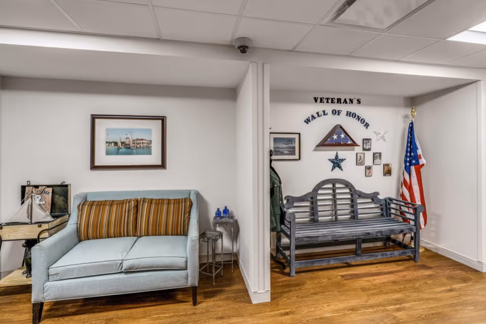 An interior space with a light blue loveseat with two striped cushions on the left, a small table with decorative items next to it, and a framed picture above. On the right, a wooden bench is placed under a wall display titled 'Veteran's Wall of Honor' featuring a folded American flag in a triangular case, stars, small framed photos, and an American flag standing in the corner. The floor is wooden and the walls are white.