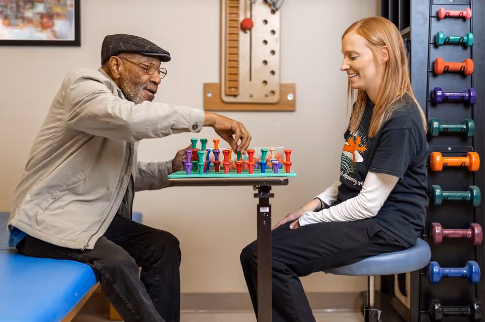 An elderly man and a young woman sitting across from each other at a small table playing a board game with colorful pegs. The man is wearing a beige jacket and a flat cap, while the woman is smiling and wearing a black shirt with a white long-sleeve underneath. Behind the woman, there is a rack with various colored dumbbells.