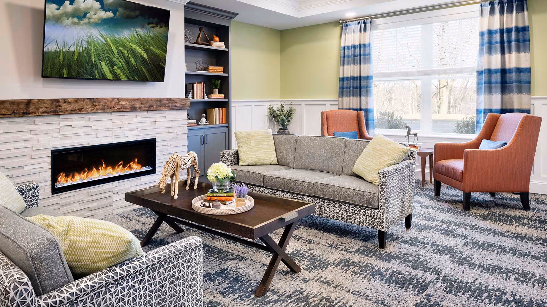 A cozy living room with a modern electric fireplace set in a white stone wall with a wooden mantel. Above the fireplace is a flat-screen TV displaying an image of green grass. The room features a gray patterned sofa with light green pillows, two orange armchairs with blue pillows, a dark wooden coffee table with decorative items, and a large window with blue and white striped curtains letting in natural light. A built-in bookshelf with books and decor is visible next to the fireplace.