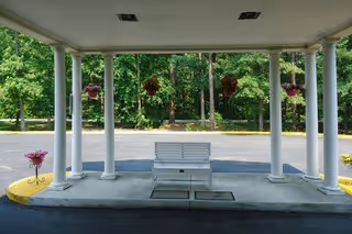 Covered entrance/porte-cochère with white columns, a white bench, hanging flower baskets, and trees beyond.