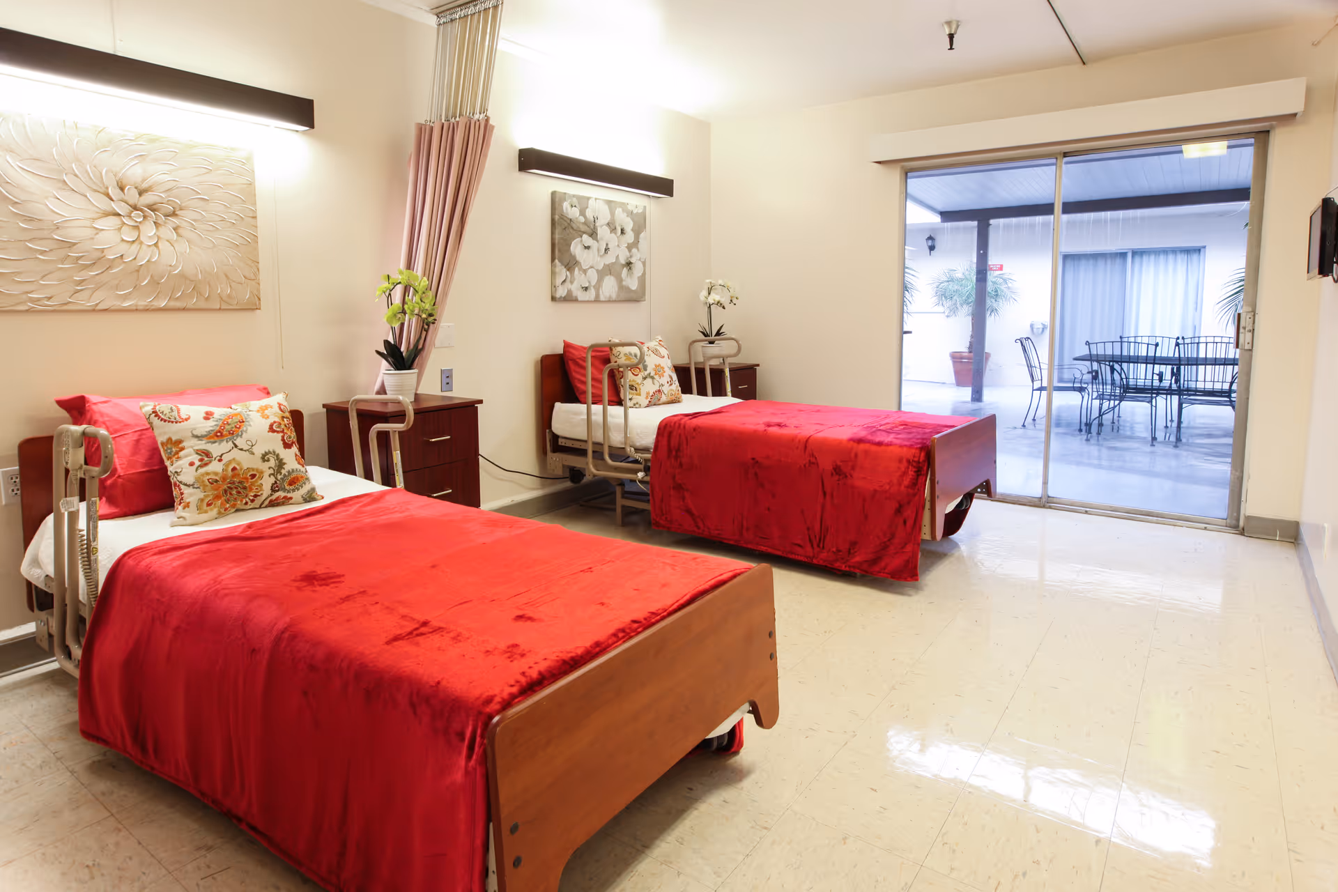 A bright room with two single beds covered in red blankets and floral pillows. Each bed has a wooden headboard and side rails. Between the beds is a small wooden nightstand with a potted plant. The walls are decorated with floral artwork and two horizontal light fixtures. A large sliding glass door opens to an outdoor patio area with a metal table and chairs.