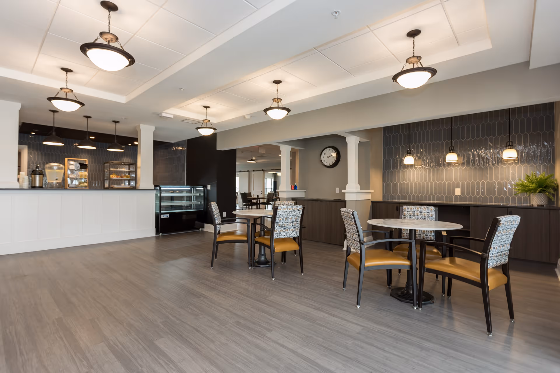 A modern dining area in a senior living facility with round tables and chairs featuring patterned backs and mustard-colored seats. The space has a light gray wood floor, pendant lights hanging from the ceiling, a clock on the wall, and a counter area with a display case and beverage dispensers in the background.