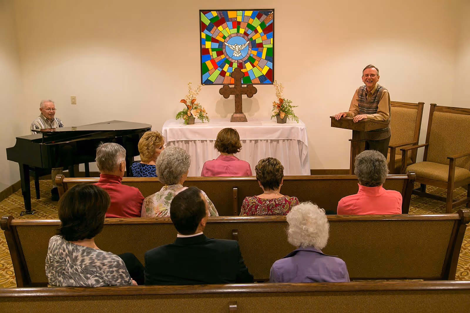 A small group of elderly people seated in pews facing a man standing at a wooden lectern in a chapel-like room. A man is playing a grand piano to the left. Behind the lectern is a table with a large wooden cross, floral arrangements, and a colorful stained glass artwork featuring a white dove.