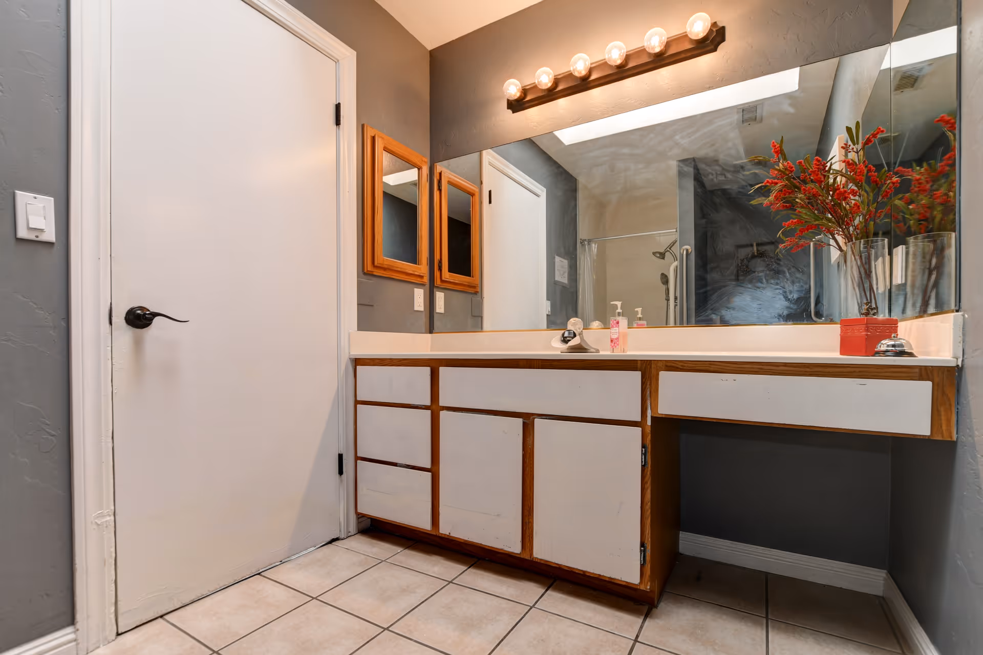 Bathroom vanity with a large mirror, multiple cabinets and drawers, a white countertop with a soap dispenser and a small bell, and a vase with red flowers. The walls are painted gray, and there is a white door to the left. Above the mirror is a light fixture with six round bulbs.