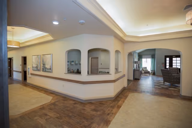 Interior view of a senior living facility hallway with light-colored walls, wood and tile flooring, and arched openings in a reception area. The hallway leads to a seating area with chairs and a window with curtains in the background.