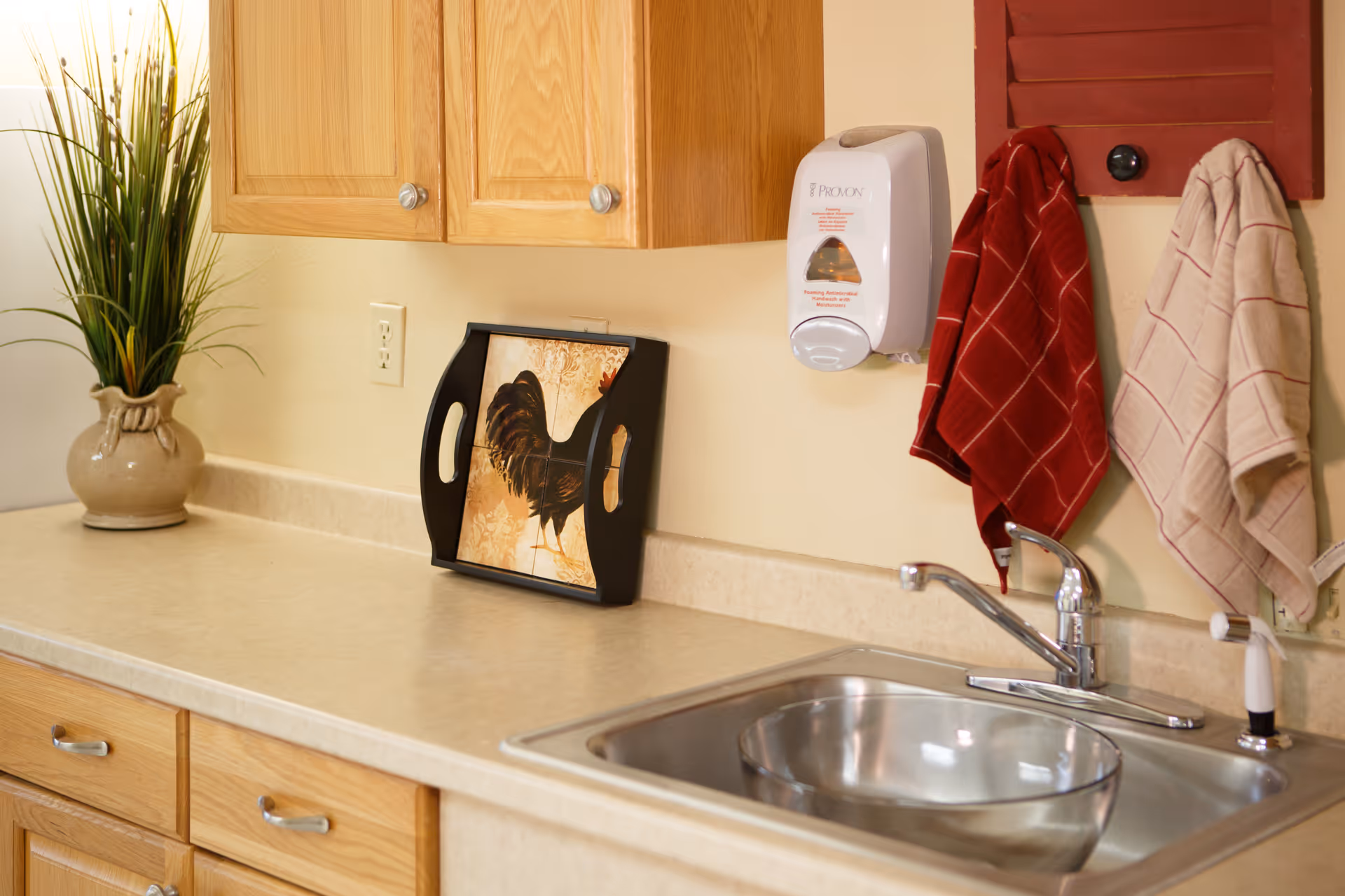 A kitchen countertop with a stainless steel sink and faucet. On the counter, there is a decorative tray with a rooster image, a beige vase with green plants, and two hanging kitchen towels, one red and one beige. Above the counter are wooden cabinets and a wall-mounted soap dispenser.