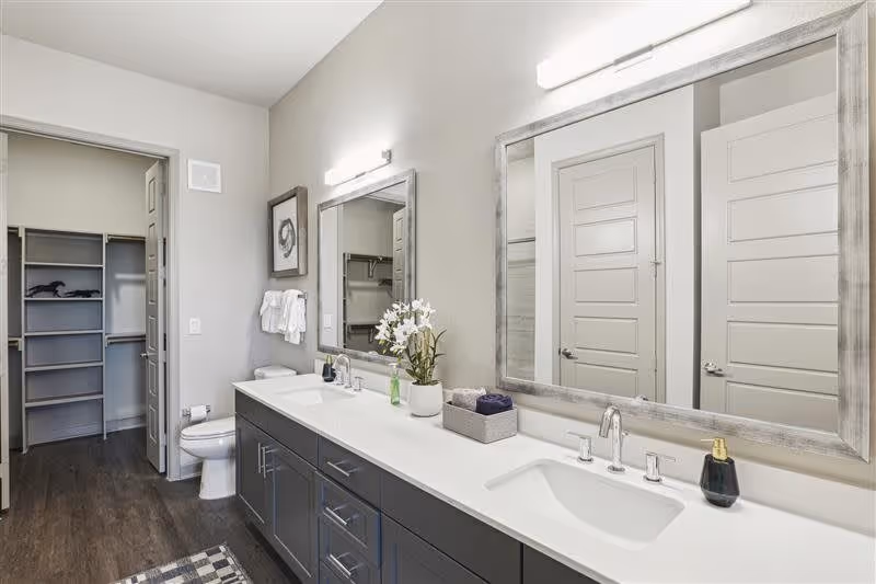Modern bathroom with a double sink vanity featuring dark cabinets and white countertops. Two large mirrors with wooden frames are mounted above the sinks, each with a light fixture above. A toilet is visible next to the vanity, and an open door reveals a walk-in closet with shelving. The floor has dark wood-style flooring and the walls are painted light gray.