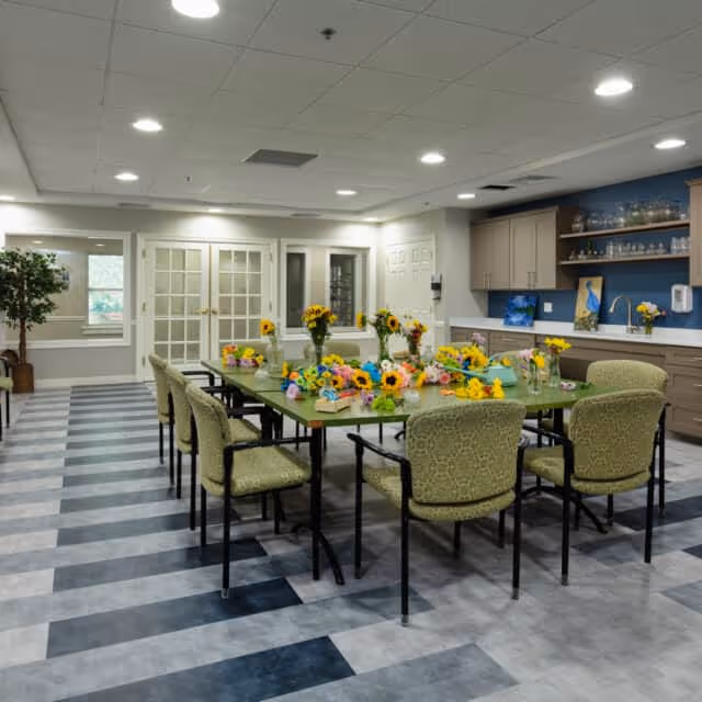 A well-lit room with a large rectangular table surrounded by eight green upholstered chairs. The table is decorated with multiple vases of colorful flowers, including sunflowers. The room has a striped floor pattern in shades of gray and blue, white walls, and a ceiling with recessed lighting. There are cabinets and shelves on one side with glassware and artwork, and double glass doors at the far end of the room.