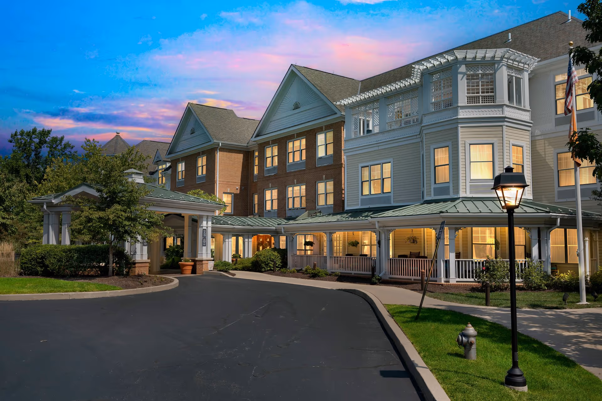 Exterior view of a senior living facility named Sunrise of Newtown Square during sunset. The building features a combination of brick and white siding with multiple windows illuminated from inside. There is a covered entrance with a driveway, well-maintained landscaping, a street lamp, and an American flag on a flagpole.