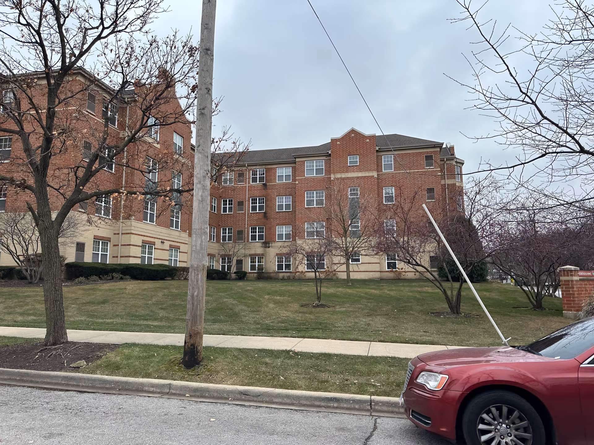 Exterior view of a multi-story brick building with multiple windows, surrounded by leafless trees and a grassy lawn. A red car is parked on the street in front of the building under a cloudy sky.
