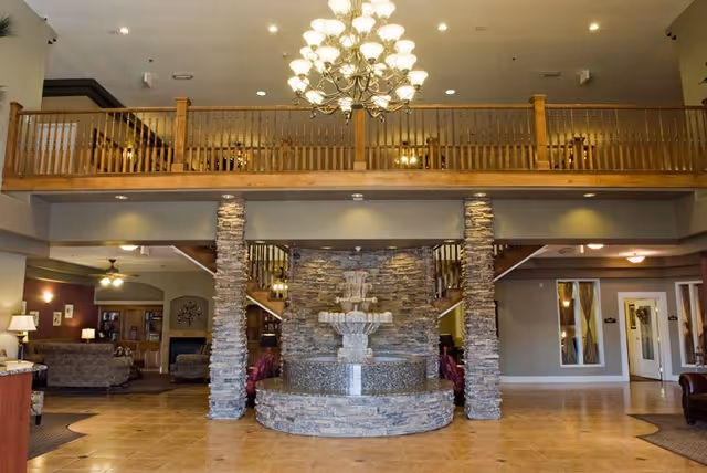 Spacious interior of a senior living facility featuring a large stone fountain in the center, flanked by two stone pillars. Above the fountain is a wooden balcony railing with a chandelier hanging from the ceiling. The area has tiled flooring and seating areas with sofas and lamps on either side.