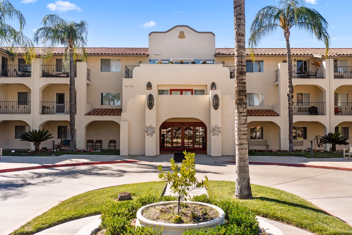 Front exterior view of a three-story senior living community building with beige stucco walls, red-tiled roof, balconies with black railings, and palm trees in front. There is a circular driveway with a small landscaped island featuring a young tree and greenery.