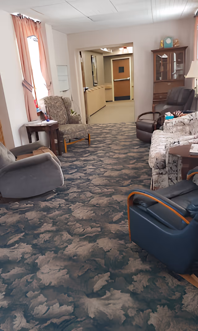 A cozy living room area in a senior living facility with various upholstered chairs and a patterned carpet. There are windows with pink curtains on the left side, a wooden side table with a lamp, and a wooden cabinet with glass doors on the right. The room opens into a hallway with a door at the end.