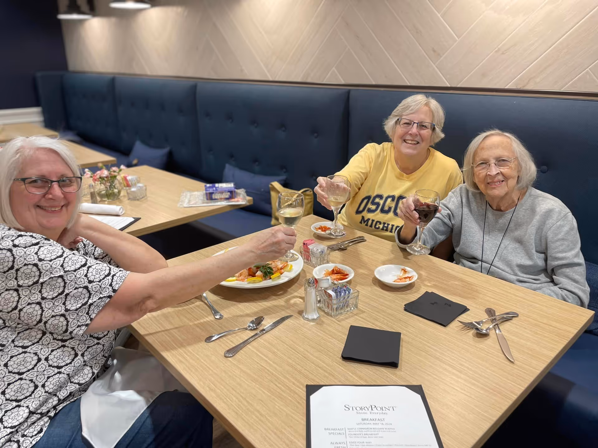 Three elderly women sitting at a dining table in a restaurant or dining area, smiling and raising their glasses in a toast. The table has plates with food, utensils, napkins, and a menu labeled StoryPoint. The seating includes a blue cushioned bench against a wall with a light-colored herringbone pattern.