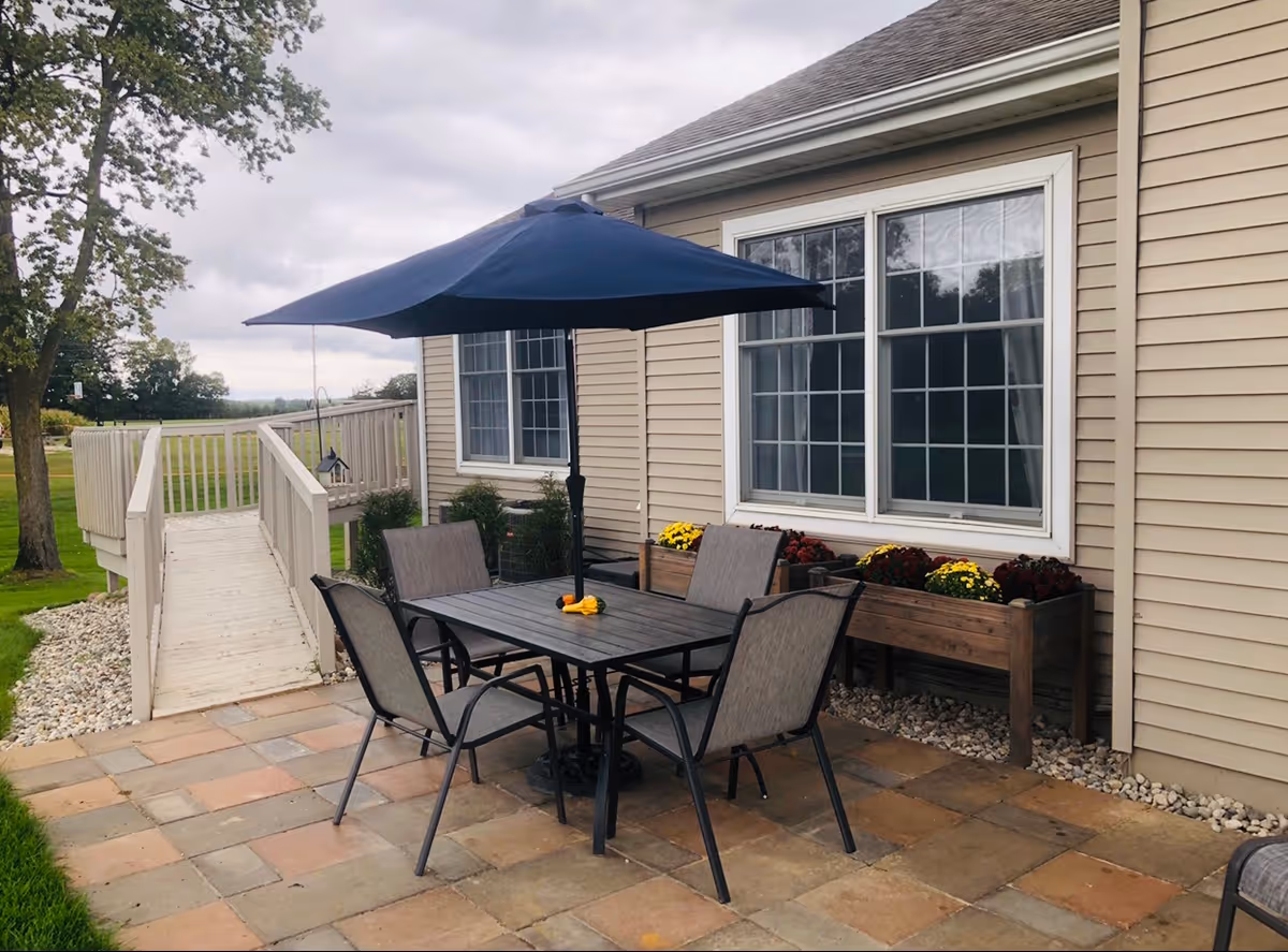 Outdoor patio area with a black metal table and four chairs under a large navy blue umbrella. The patio is paved with square tiles and is adjacent to a beige building with two large windows and flower boxes filled with colorful flowers. A wooden ramp with railings leads away from the patio towards a grassy area with trees.