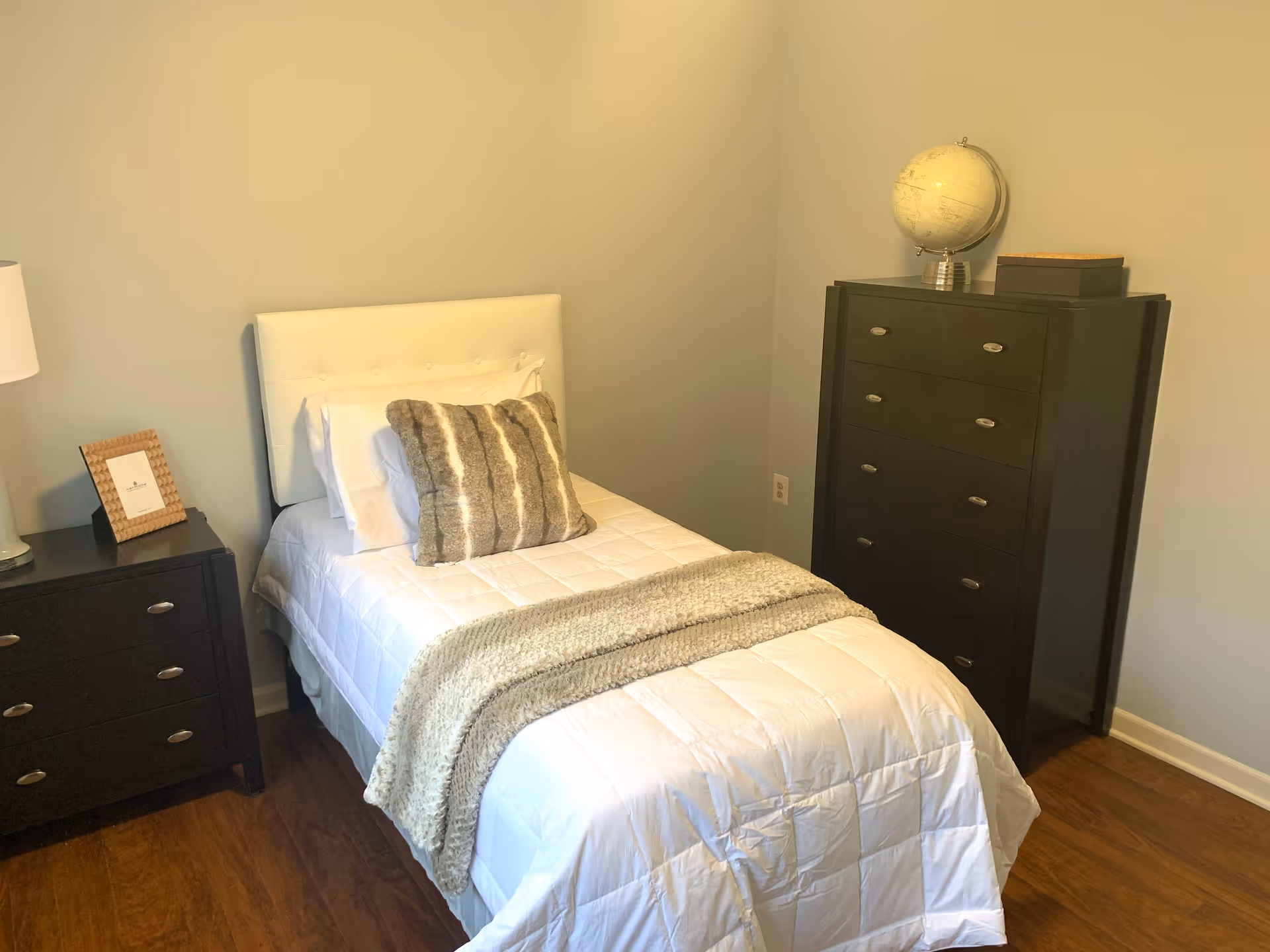 A small bedroom with a single bed made with white bedding, a gray and white striped pillow, and a gray throw blanket. To the left of the bed is a dark wooden nightstand with a lamp and a framed picture. To the right of the bed is a tall dark wooden dresser with a globe and a small box on top. The room has light-colored walls and wooden flooring.